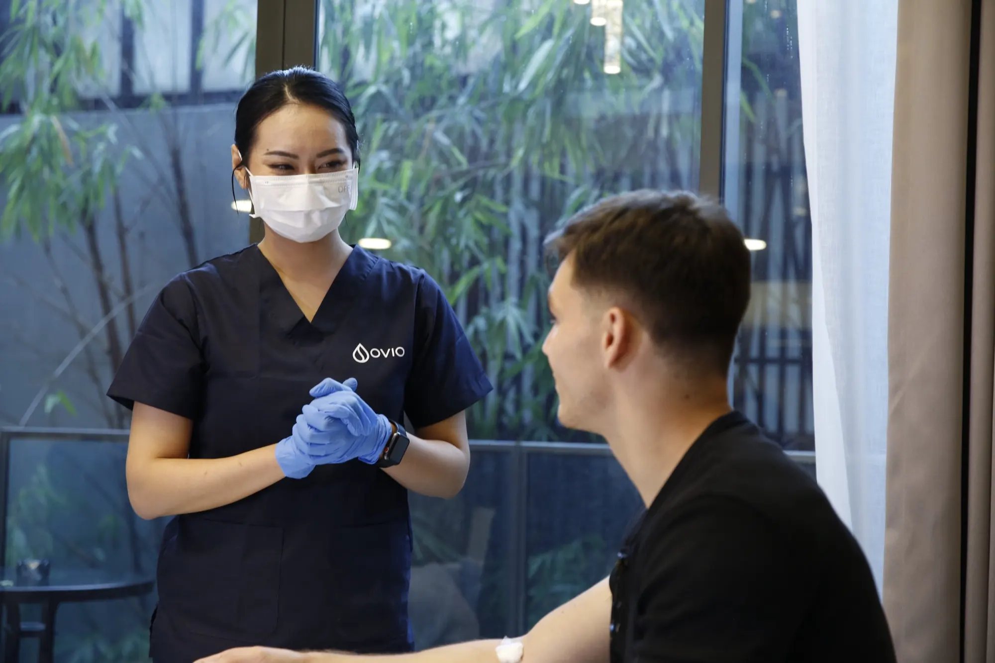 A smiling Ovio nurse talking with a client after his blood draw, a cotton pad taped to his arm, in a Bangkok condo