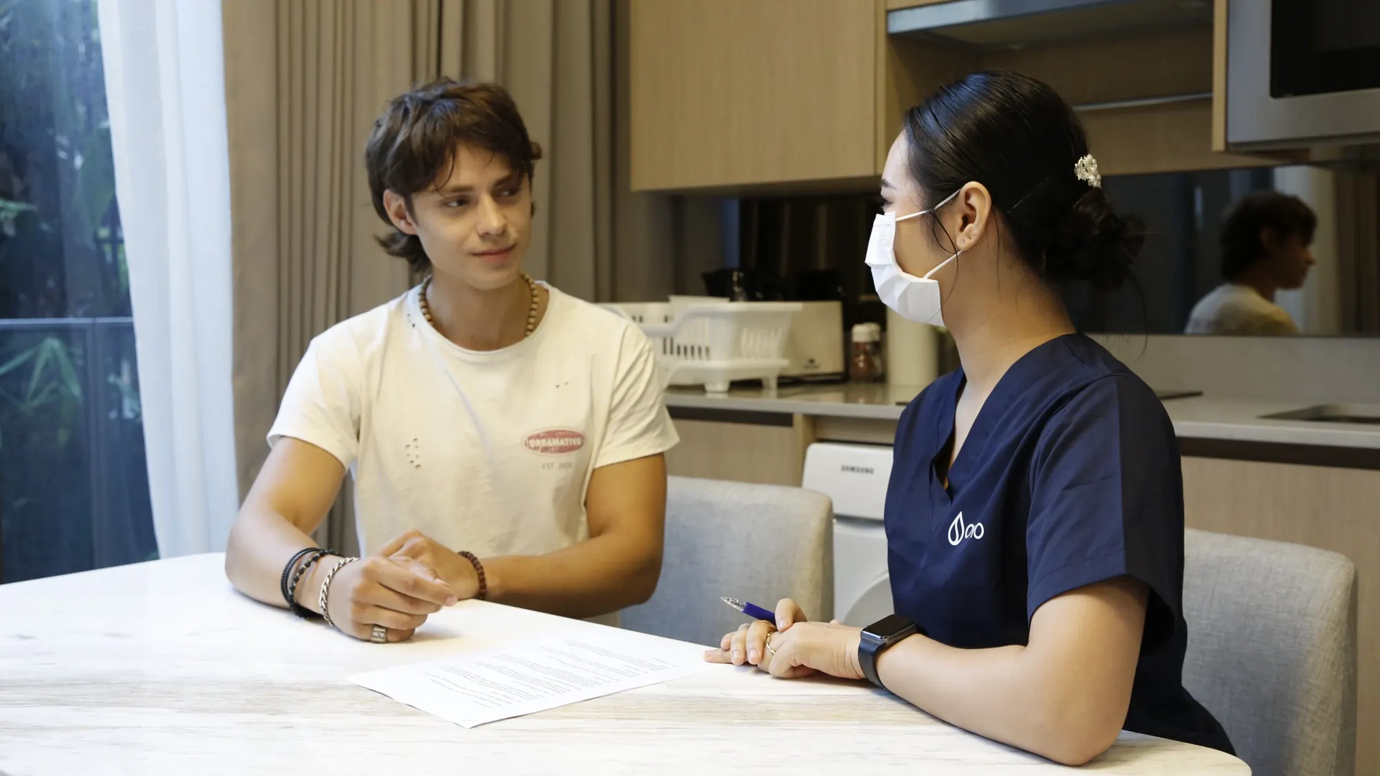 An Ovio nurse talking privately with a client at his kitchen table in a Bangkok condo before his test
